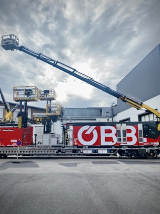 A railway maintenance vehicle with large red and white branding on its side is parked on a track. It is equipped with several mechanical arms and platforms, likely used for maintenance tasks. Above, the sky is overcast with clouds.