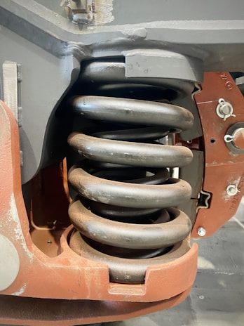 A close-up view of a large metal coil spring integrated into a mechanical assembly. The spring is encased in a sturdy, rust-colored metal frame which supports its structure, featuring several fasteners and joints. The metal has a matte finish and signs of wear, suggesting industrial use.
