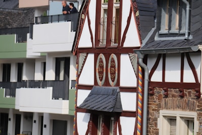 A half-timbered house with ornate design and wooden beams stands next to a modern apartment building with balconies. Two people look out from one of the balconies.