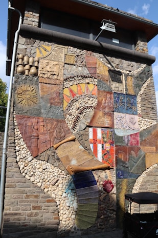 A vibrant and intricate mural displayed on an exterior wall, composed of various textures and materials such as stone, ceramic, and metal. It features an artistic collage of symbols, patterns, and reliefs depicting historical themes and abstract designs. The mural includes sun motifs, coats of arms, and scrolls with inscriptions.