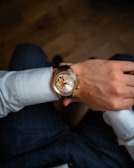 A close-up view of a person's wrist adorned with an elegant wristwatch featuring a transparent face that reveals intricate gears and mechanical components. The attire includes a white dress shirt and dark pinstripe trousers, suggesting a formal or business setting.