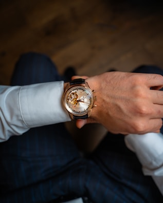 A close-up view of a person's wrist adorned with an elegant wristwatch featuring a transparent face that reveals intricate gears and mechanical components. The attire includes a white dress shirt and dark pinstripe trousers, suggesting a formal or business setting.