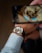A close-up view of a wristwatch being worn on a person's wrist. The watch has a gold bezel, a silver face with visible gears, and is attached to a black leather strap. A smartphone appears to be capturing an image of the watch, with part of the phone visible in the frame.