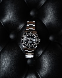 A luxury watch with a stainless steel bracelet and black dial is prominently displayed on a textured black leather surface. The watch face features luminous hour markers, a date window, and a rotating bezel marked with numerals.