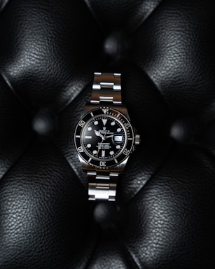 A luxury watch with a stainless steel bracelet and black dial is prominently displayed on a textured black leather surface. The watch face features luminous hour markers, a date window, and a rotating bezel marked with numerals.
