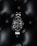 A luxury watch with a stainless steel bracelet and black dial is prominently displayed on a textured black leather surface. The watch face features luminous hour markers, a date window, and a rotating bezel marked with numerals.