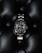 A luxury watch with a stainless steel bracelet and black dial is prominently displayed on a textured black leather surface. The watch face features luminous hour markers, a date window, and a rotating bezel marked with numerals.