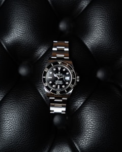 A luxury watch with a stainless steel bracelet and black dial is prominently displayed on a textured black leather surface. The watch face features luminous hour markers, a date window, and a rotating bezel marked with numerals.