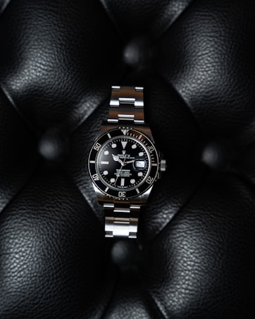 A luxury watch with a stainless steel bracelet and black dial is prominently displayed on a textured black leather surface. The watch face features luminous hour markers, a date window, and a rotating bezel marked with numerals.