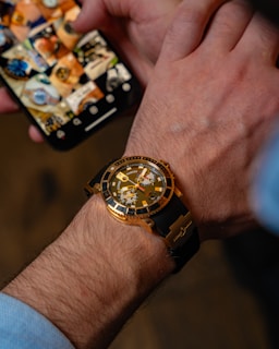 A close-up of a person's wrist wearing a gold and black watch. The watch features a large dial with visible markings and hands. In the background, a smartphone displays a grid of images, possibly of various watches.
