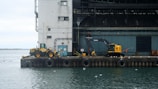 An industrial port scene with construction equipment stands on a concrete pier against a large industrial building. The overcast sky casts a muted light over the scene, and numerous seagulls are scattered across the pier and the water. The excavator and loader suggest ongoing or recent construction activities.