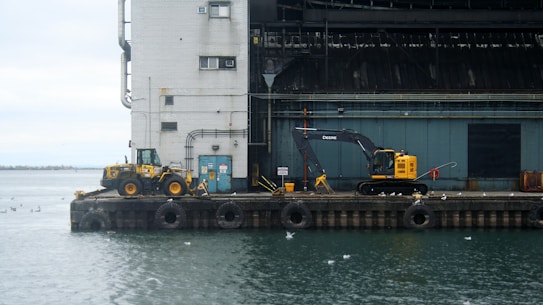 An industrial port scene with construction equipment stands on a concrete pier against a large industrial building. The overcast sky casts a muted light over the scene, and numerous seagulls are scattered across the pier and the water. The excavator and loader suggest ongoing or recent construction activities.