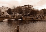 A river runs through a city scene with various buildings in the background, including an advertisement for Minerva Automobielen. Trees line the riverbank, and several small boats are moored along the edge. The architecture reflects an urban environment with a touch of greenery.