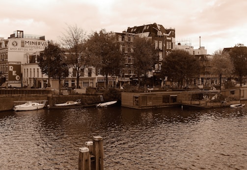 A river runs through a city scene with various buildings in the background, including an advertisement for Minerva Automobielen. Trees line the riverbank, and several small boats are moored along the edge. The architecture reflects an urban environment with a touch of greenery.