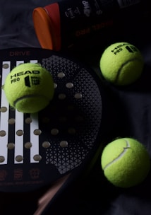 A padel racket lies on a dark surface, accompanied by three bright yellow padel balls. The balls are scattered around the racket, which has a circular grid design with large holes. A cylindrical container with an orange lid, likely a ball canister, is partially visible.