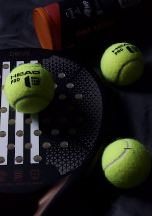 A padel racket lies on a dark surface, accompanied by three bright yellow padel balls. The balls are scattered around the racket, which has a circular grid design with large holes. A cylindrical container with an orange lid, likely a ball canister, is partially visible.