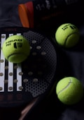 A padel racket lies on a dark surface, accompanied by three bright yellow padel balls. The balls are scattered around the racket, which has a circular grid design with large holes. A cylindrical container with an orange lid, likely a ball canister, is partially visible.