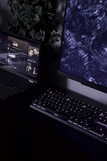 A dimly lit workspace containing a partially visible laptop displaying a music streaming service with an album cover titled 'chill', a mechanical keyboard with backlit keys, and a monitor displaying abstract dark patterns. In the background, there is a plant with green leaves adding a natural element to the setup.