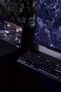 A dimly lit workspace containing a partially visible laptop displaying a music streaming service with an album cover titled 'chill', a mechanical keyboard with backlit keys, and a monitor displaying abstract dark patterns. In the background, there is a plant with green leaves adding a natural element to the setup.