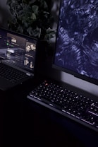 A dimly lit workspace containing a partially visible laptop displaying a music streaming service with an album cover titled 'chill', a mechanical keyboard with backlit keys, and a monitor displaying abstract dark patterns. In the background, there is a plant with green leaves adding a natural element to the setup.