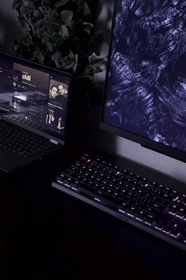 A dimly lit workspace containing a partially visible laptop displaying a music streaming service with an album cover titled 'chill', a mechanical keyboard with backlit keys, and a monitor displaying abstract dark patterns. In the background, there is a plant with green leaves adding a natural element to the setup.