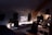 A stylish and modern computer setup inside what appears to be a cozy attic. The setup features a large monitor displaying an ocean scene, a keyboard, and a mouse on a sleek black desk. Decorative plants and ambient lighting give the space a serene and natural feel. There is a comfortable chair in front of the desk, and a computer tower is situated next to the monitor. Some shelves and a framed picture adorn the wall.