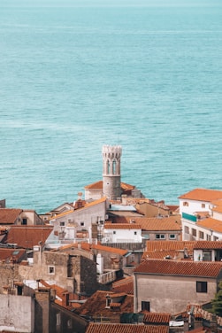 A coastal townscape with a prominent white clock tower rising above an array of terracotta-roofed buildings. The expansive teal ocean stretches in the background.