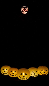 A mysterious scene featuring a dark background with several illuminated pumpkin lanterns forming a row at the bottom of the image. Above, a glowing mask with a sinister smile and stitched eyes floats in the darkness, enhancing the eerie atmosphere.