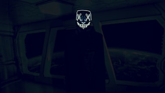 A dark, futuristic room with an individual wearing a hooded cloak and a glowing mask with crossed eyes and a stitched mouth design. The background features large windows displaying a view of Earth from space, creating an eerie and mysterious atmosphere.