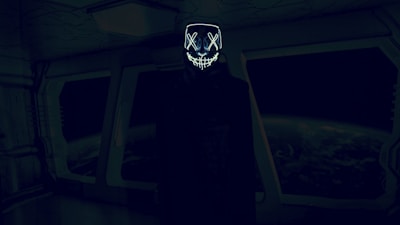 A dark, futuristic room with an individual wearing a hooded cloak and a glowing mask with crossed eyes and a stitched mouth design. The background features large windows displaying a view of Earth from space, creating an eerie and mysterious atmosphere.
