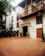 A charming courtyard featuring terracotta-tiled flooring, surrounded by a two-story building with wooden balconies adorned with potted plants. The white facade contrasts with the rich brown woodwork. Multiple black lampposts line the walkway, and leafy green plants provide a touch of nature. The entrance to a restaurant or bar named 'Alma' is visible, framed by a stone archway.
