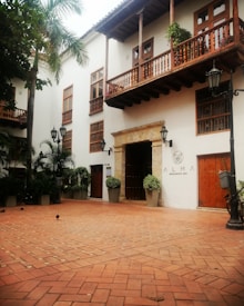 A charming courtyard featuring terracotta-tiled flooring, surrounded by a two-story building with wooden balconies adorned with potted plants. The white facade contrasts with the rich brown woodwork. Multiple black lampposts line the walkway, and leafy green plants provide a touch of nature. The entrance to a restaurant or bar named 'Alma' is visible, framed by a stone archway.
