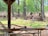 A collection of old, rusty farming machinery is scattered in a grassy area surrounded by tall trees. These machines have large metal wheels and are mostly covered with rust, indicating they are likely antique. A wooden table is partially visible in the foreground, situated under a wooden structure.