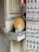 An orange tabby cat is drinking water from a glass placed on a stone ledge. The cat is positioned next to a display of postcards featuring historical landmarks. The scene appears to be outside a store with a weathered stone wall.
