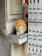 An orange tabby cat is drinking water from a glass placed on a stone ledge. The cat is positioned next to a display of postcards featuring historical landmarks. The scene appears to be outside a store with a weathered stone wall.