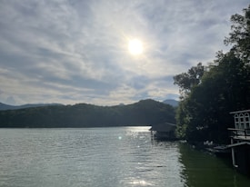 A serene lakeside view with the sun shining above a slightly cloudy sky. The water reflects the sunlight, creating a sparkling effect. On the right, there are trees and a boathouse extending over the water.