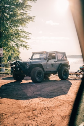 A rugged off-road vehicle is parked on a dirt path near a body of water, with sunlight casting strong shadows. The vehicle's large tires and sturdy build suggest it's made for rough terrain. Trees provide a natural backdrop, and the water glistens under the bright sun.
