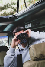 A close-up of a safari backpack with binoculars and a hat resting on a jeep dashboard at sunrise.