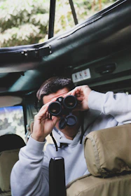 A close-up of a safari backpack with binoculars and a hat resting on a jeep dashboard at sunrise.