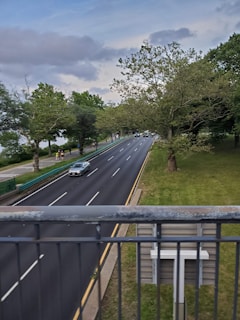 A road with several lanes is bordered by lush green trees on one side and a metal railing in the foreground. There are a few vehicles on the road, including a silver car, traveling along the freshly paved highway. The sky is partly cloudy, casting a calm atmosphere over the scene.