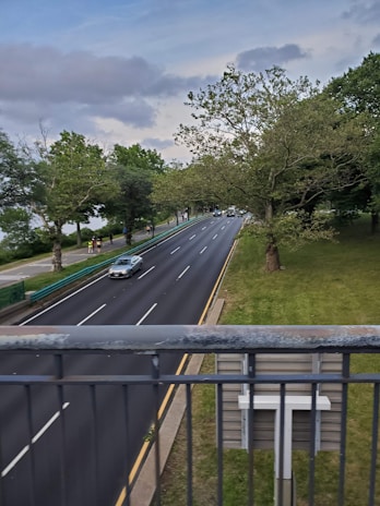A road with several lanes is bordered by lush green trees on one side and a metal railing in the foreground. There are a few vehicles on the road, including a silver car, traveling along the freshly paved highway. The sky is partly cloudy, casting a calm atmosphere over the scene.