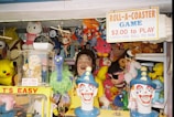 A carnival game booth filled with various colorful plush toys and figures. A sign on top advertises a game costing $2.00 to play, with a challenge to catch a ball to win. The booth is decorated with well-known character plushies, including a Pikachu, Hello Kitty, and Shrek. Two clown heads are prominently displayed in the foreground, painted with bright colors and exaggerated features.