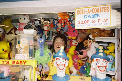 A carnival game booth filled with various colorful plush toys and figures. A sign on top advertises a game costing $2.00 to play, with a challenge to catch a ball to win. The booth is decorated with well-known character plushies, including a Pikachu, Hello Kitty, and Shrek. Two clown heads are prominently displayed in the foreground, painted with bright colors and exaggerated features.