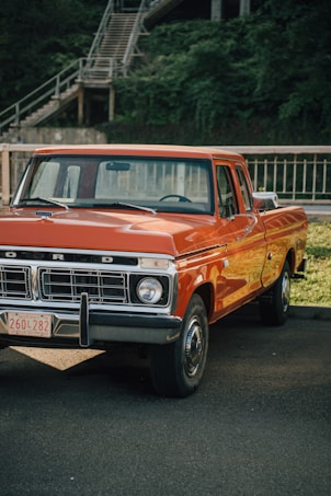 An orange vintage pickup truck is parked on a paved area with green foliage and a staircase in the background. The truck's front features a silver grille and round headlights, with a Quebec license plate.
