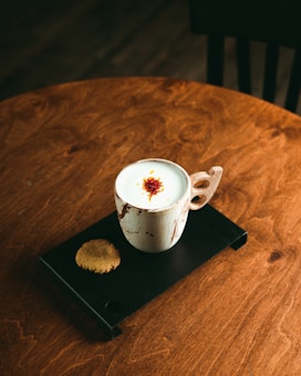 A rustic wooden table holds a black serving tray with a white, speckled ceramic mug containing a frothy beverage topped with saffron strands. Accompanying the mug is a round cookie, adding an element of warmth to the cozy setting.