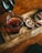 A glass filled with a dark red liquid being poured from a pot. The setup includes a wooden tray with slices of dried citrus, a cinnamon stick, and a chocolate-coated treat on a stick. The overall ambiance is warm and rustic.