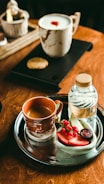 A wooden table is set with a metal tray containing a brown mug of coffee, a bottle of water, and a selection of sliced fruits including strawberries and grapes. Nearby, a light-colored ceramic mug with a frothy beverage and a cookie on a black tray completes the cozy setting.