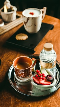 A wooden table is set with a metal tray containing a brown mug of coffee, a bottle of water, and a selection of sliced fruits including strawberries and grapes. Nearby, a light-colored ceramic mug with a frothy beverage and a cookie on a black tray completes the cozy setting.