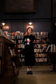 A woman stands in the center of a dimly lit library, surrounded by shelves filled with books. She is dressed in a black outfit, holding a book in her hand, and looks thoughtfully to the side. The warm lighting creates a cozy and contemplative atmosphere.
