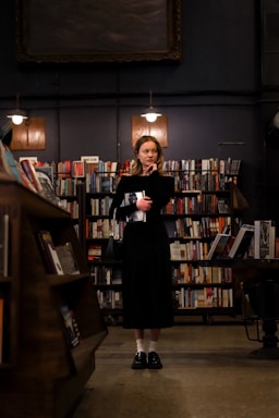 A woman stands in the center of a dimly lit library, surrounded by shelves filled with books. She is dressed in a black outfit, holding a book in her hand, and looks thoughtfully to the side. The warm lighting creates a cozy and contemplative atmosphere.